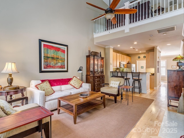 living area with crown molding, light wood-type flooring, a high ceiling, ceiling fan, and visible vents