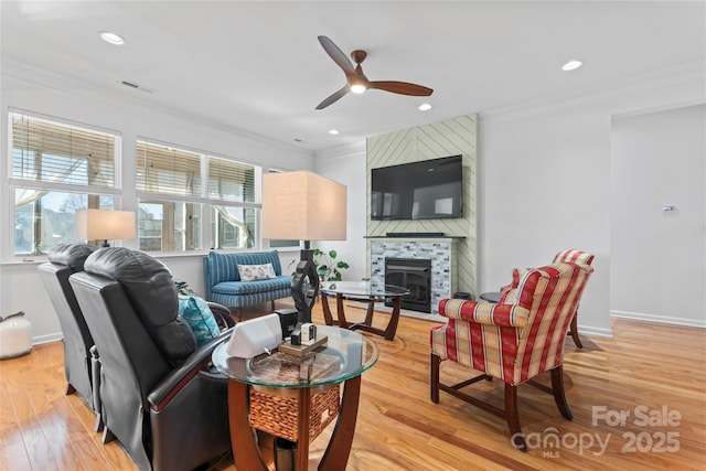 living area featuring light wood finished floors, baseboards, ornamental molding, a fireplace, and a ceiling fan