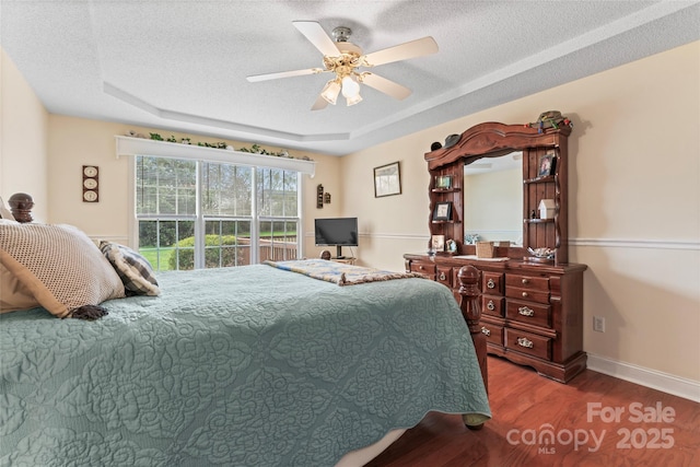 bedroom with a tray ceiling, a textured ceiling, wood finished floors, baseboards, and ceiling fan