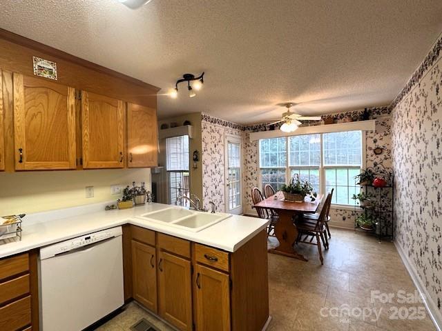 kitchen featuring brown cabinetry, wallpapered walls, a peninsula, white dishwasher, and a sink