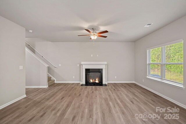 unfurnished living room with stairway, wood finished floors, a ceiling fan, visible vents, and a fireplace with flush hearth