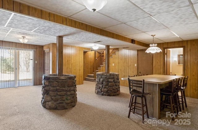 interior space with a drop ceiling, carpet, a bar, wood walls, and stairs