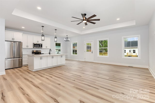 unfurnished living room with ceiling fan with notable chandelier, a tray ceiling, light wood-style flooring, recessed lighting, and baseboards