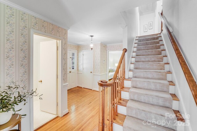 stairs with crown molding, wainscoting, wallpapered walls, and wood finished floors