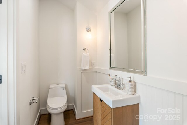 half bath featuring vanity, toilet, wood finished floors, and wainscoting