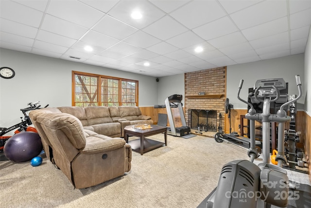 carpeted living room with a drop ceiling, a wainscoted wall, wooden walls, visible vents, and a brick fireplace