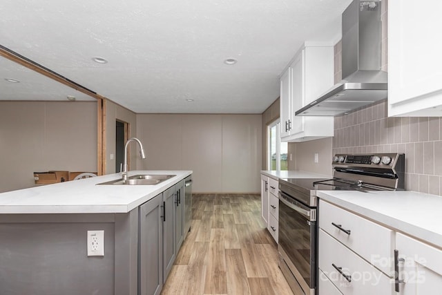 kitchen with appliances with stainless steel finishes, wall chimney exhaust hood, a center island with sink, light wood-style floors, and a sink