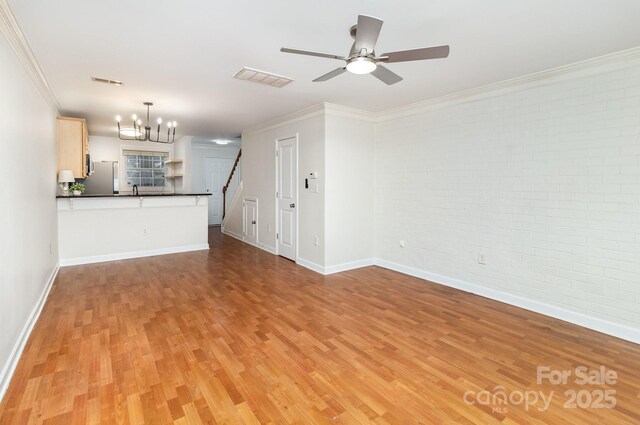 unfurnished living room with visible vents, brick wall, ornamental molding, light wood finished floors, and ceiling fan with notable chandelier