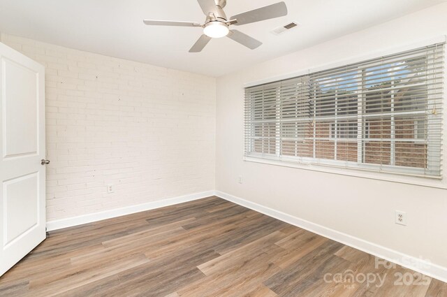 unfurnished room featuring ceiling fan, visible vents, baseboards, brick wall, and wood finished floors