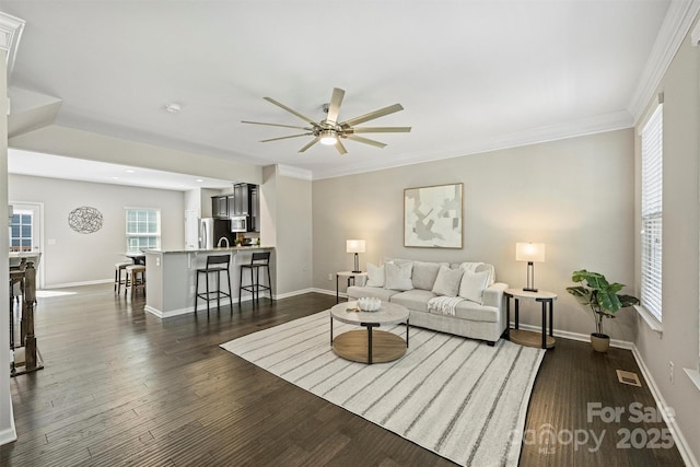 living area with dark wood-type flooring, crown molding, baseboards, and ceiling fan