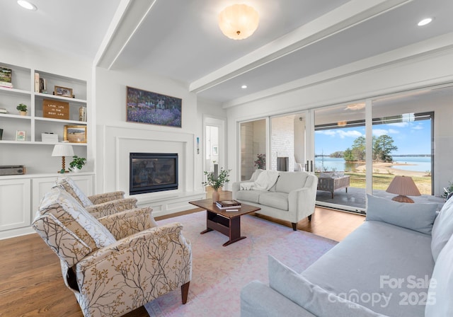 living area with wood finished floors, a glass covered fireplace, a water view, beamed ceiling, and recessed lighting