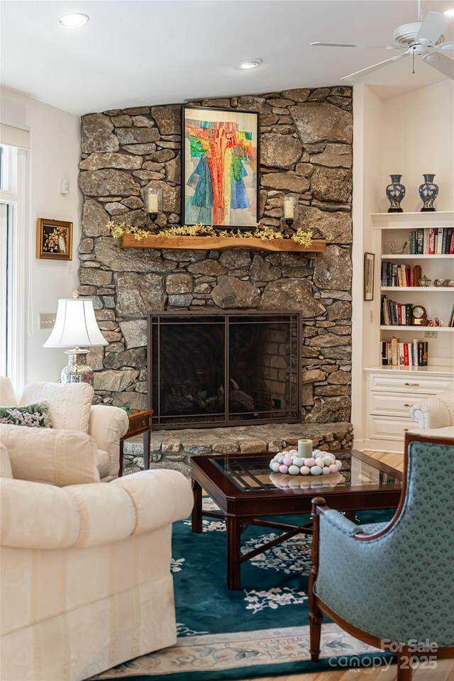 living room featuring a stone fireplace, built in shelves, and ceiling fan