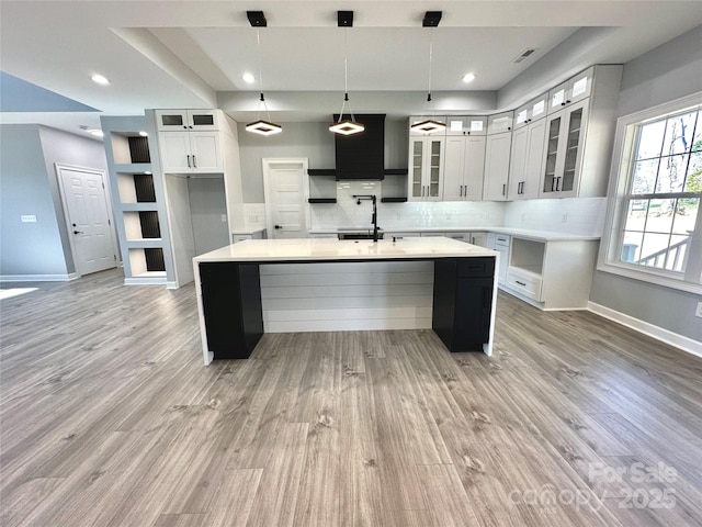 kitchen featuring an island with sink, backsplash, white cabinets, light wood finished floors, and baseboards