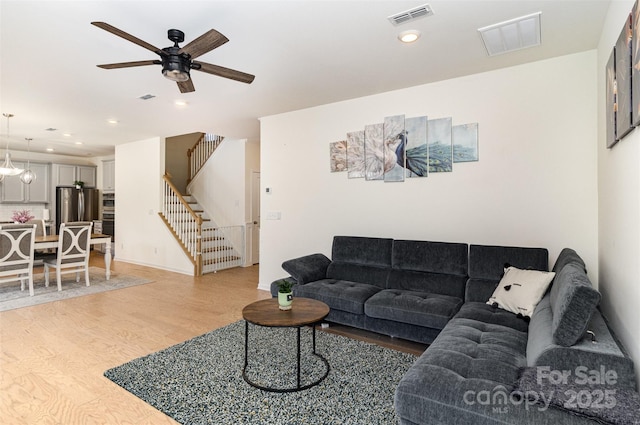 living room featuring light wood-type flooring, ceiling fan, visible vents, and stairway