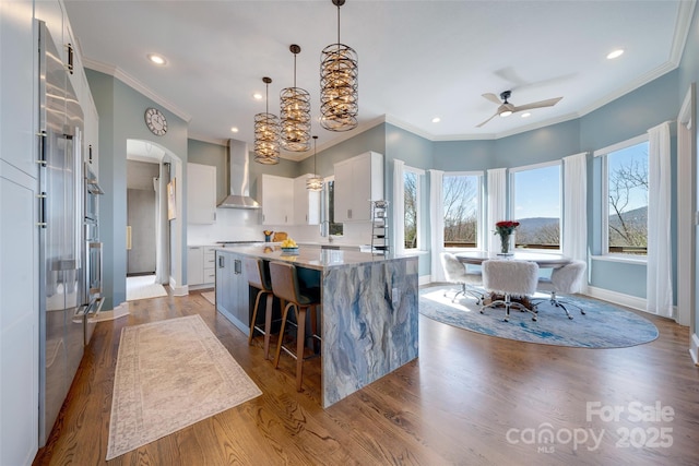 kitchen with light wood-style flooring, a breakfast bar, wall chimney exhaust hood, arched walkways, and ceiling fan with notable chandelier