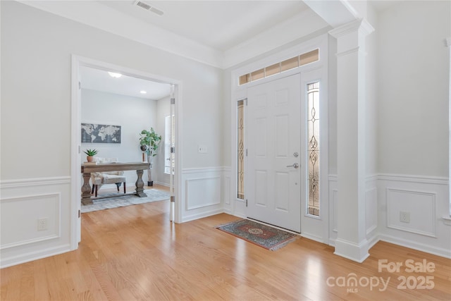 foyer entrance with light wood-style floors, a decorative wall, decorative columns, visible vents, and wainscoting
