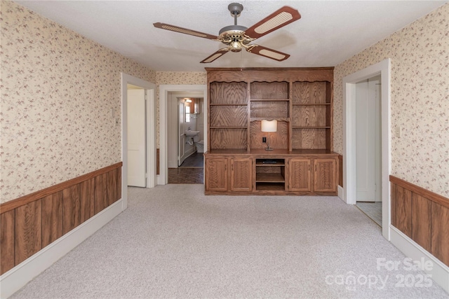 unfurnished living room with wainscoting, ceiling fan, carpet flooring, and wallpapered walls