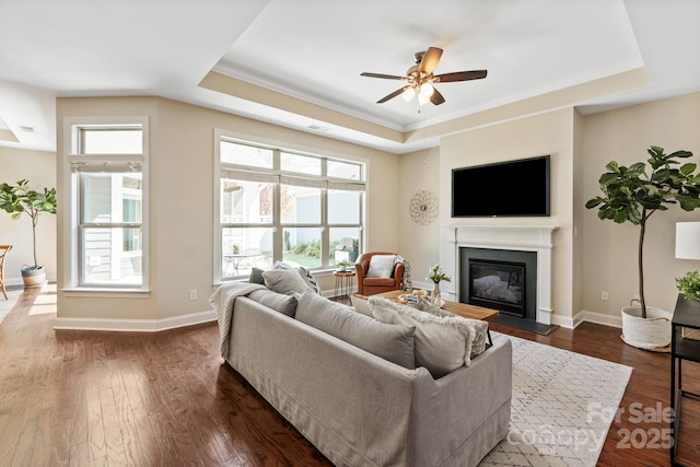 living room featuring dark wood-style flooring, baseboards, ceiling fan, a raised ceiling, and a glass covered fireplace