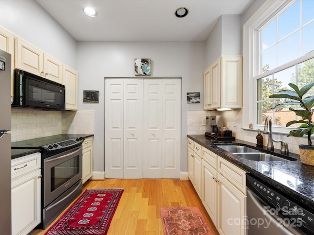 kitchen with a sink, appliances with stainless steel finishes, light wood-type flooring, cream cabinets, and dark stone countertops