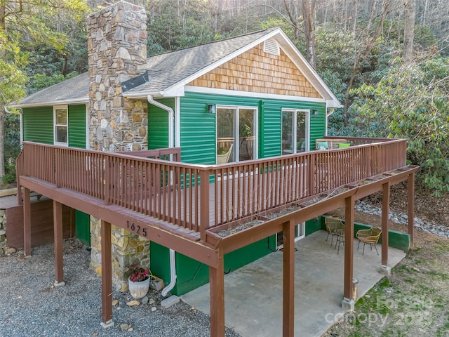 rear view of house featuring stone siding, a view of trees, a wooden deck, a shingled roof, and a patio