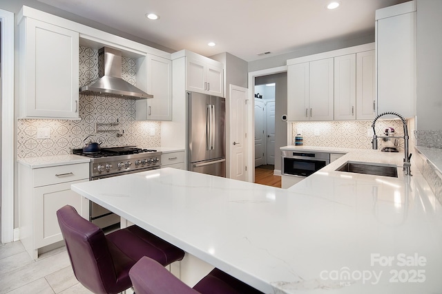 kitchen featuring a sink, a breakfast bar area, light stone countertops, appliances with stainless steel finishes, and wall chimney range hood