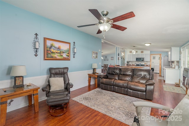 living area featuring a ceiling fan, a wainscoted wall, and dark wood-type flooring