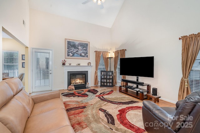 living area with high vaulted ceiling, a glass covered fireplace, baseboards, wood finished floors, and ceiling fan