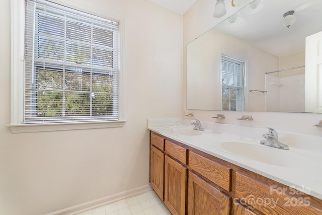 bathroom featuring a shower, double vanity, a sink, and baseboards