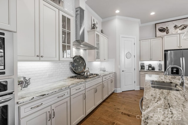 kitchen featuring a sink, wood finished floors, appliances with stainless steel finishes, wall chimney range hood, and ornamental molding