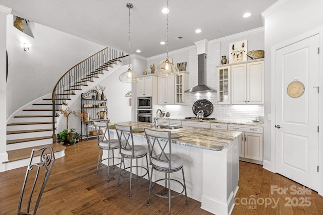 kitchen featuring wall chimney exhaust hood, a center island with sink, dark wood finished floors, a sink, and a breakfast bar area