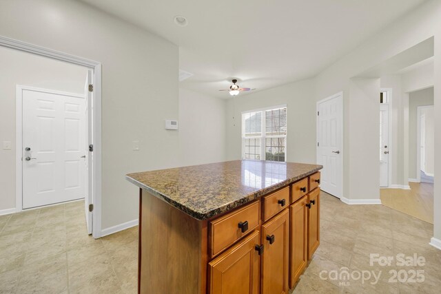 kitchen featuring a ceiling fan, a center island, brown cabinets, dark stone countertops, and baseboards