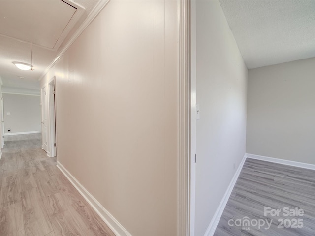 hallway featuring attic access, light wood-type flooring, ornamental molding, baseboards, and a textured ceiling