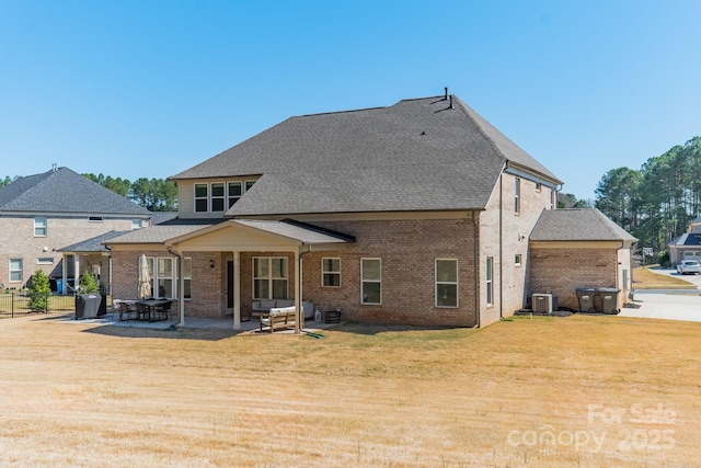 rear view of property with brick siding, roof with shingles, a lawn, cooling unit, and a patio area