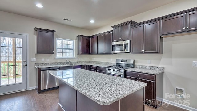 kitchen featuring light stone counters, dark brown cabinetry, a sink, a kitchen island, and appliances with stainless steel finishes