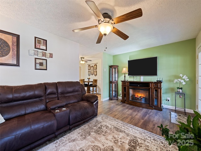 living area featuring a textured ceiling, a glass covered fireplace, a ceiling fan, baseboards, and wood finished floors