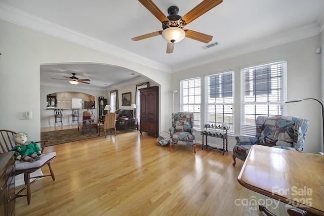 living area featuring light wood-style floors, visible vents, arched walkways, ceiling fan, and ornamental molding