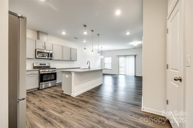 kitchen with dark wood-style flooring, a sink, a kitchen island with sink, tasteful backsplash, and appliances with stainless steel finishes