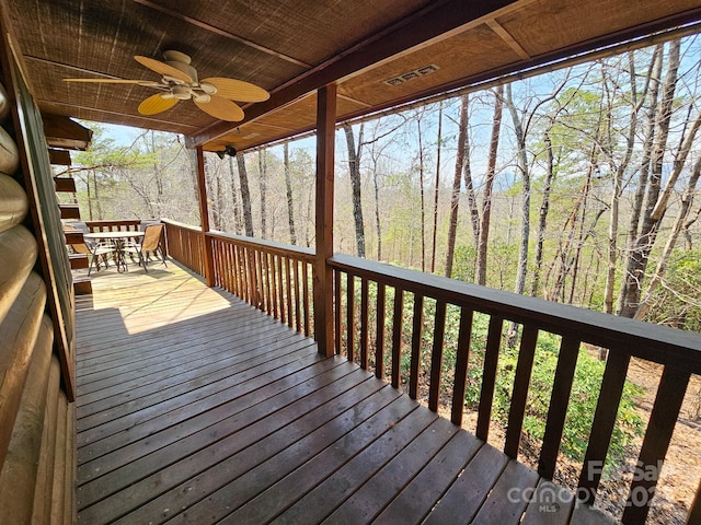 wooden terrace with a wooded view, outdoor dining area, ceiling fan, and visible vents