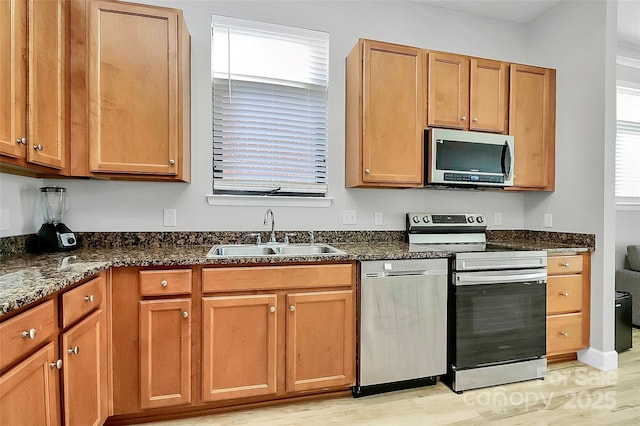 kitchen with brown cabinets, dark stone counters, stainless steel appliances, a sink, and light wood-type flooring