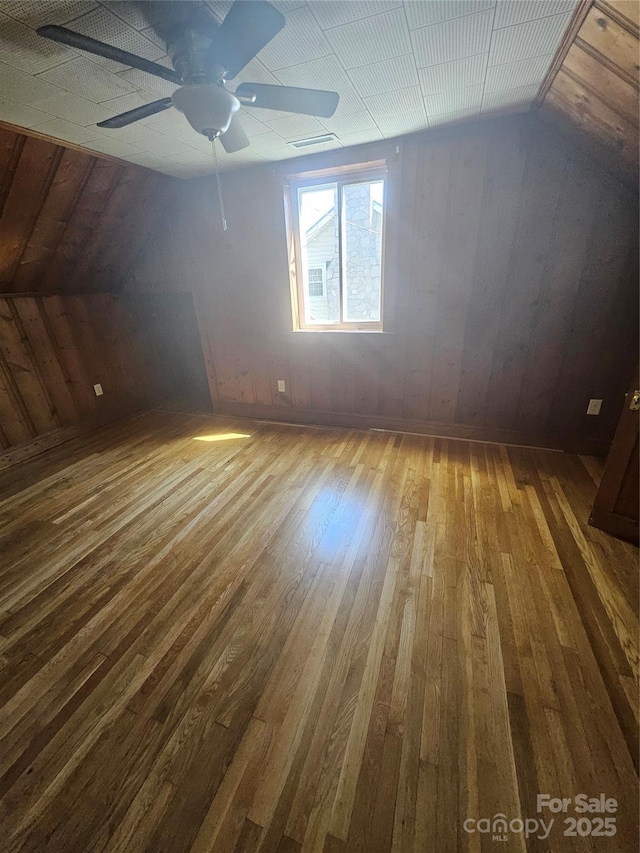 additional living space with wood walls, lofted ceiling, hardwood / wood-style flooring, visible vents, and a ceiling fan