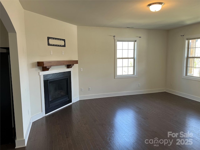 unfurnished living room featuring arched walkways, a glass covered fireplace, baseboards, and dark wood finished floors