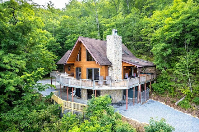 back of property featuring a wooden deck, a wooded view, a chimney, and stone siding