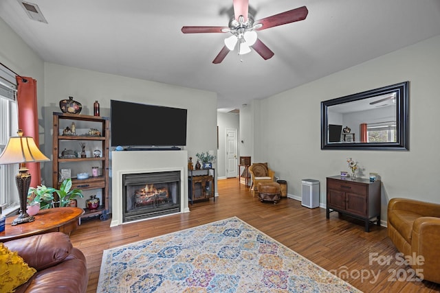 living room with a lit fireplace, a ceiling fan, wood finished floors, baseboards, and visible vents