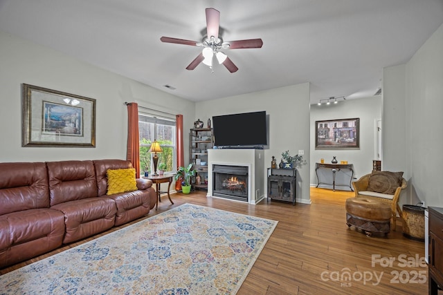living room featuring a warm lit fireplace, visible vents, baseboards, ceiling fan, and wood finished floors