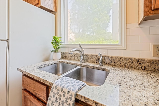 kitchen featuring a sink, freestanding refrigerator, brown cabinetry, tasteful backsplash, and light stone counters
