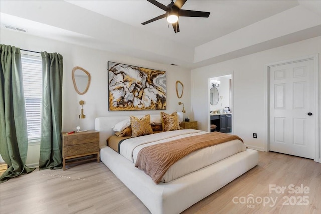 bedroom with a raised ceiling, light wood-style flooring, ensuite bathroom, baseboards, and visible vents