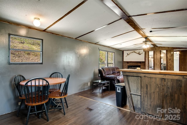 dining space with lofted ceiling with beams, dark wood-style floors, a ceiling fan, and a brick fireplace