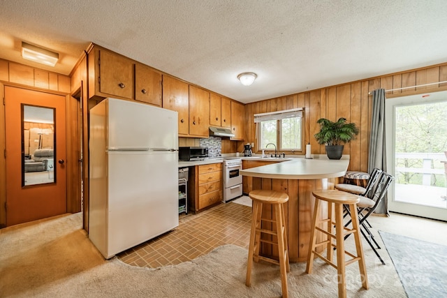 kitchen featuring a peninsula, wood walls, white appliances, light countertops, and under cabinet range hood