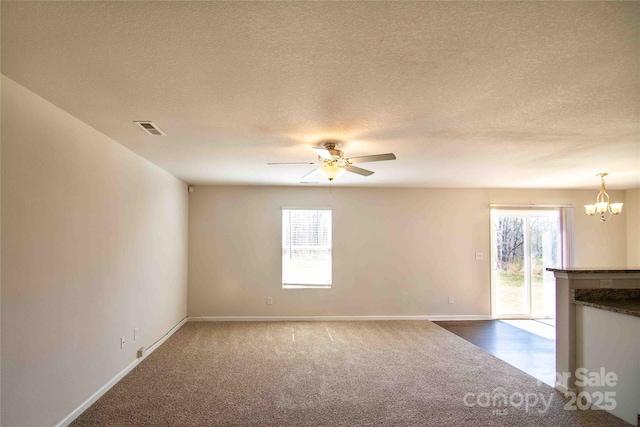 unfurnished living room with visible vents, dark colored carpet, ceiling fan with notable chandelier, and plenty of natural light