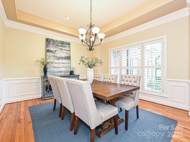 dining room featuring a raised ceiling, light wood-type flooring, a chandelier, ornamental molding, and a wainscoted wall